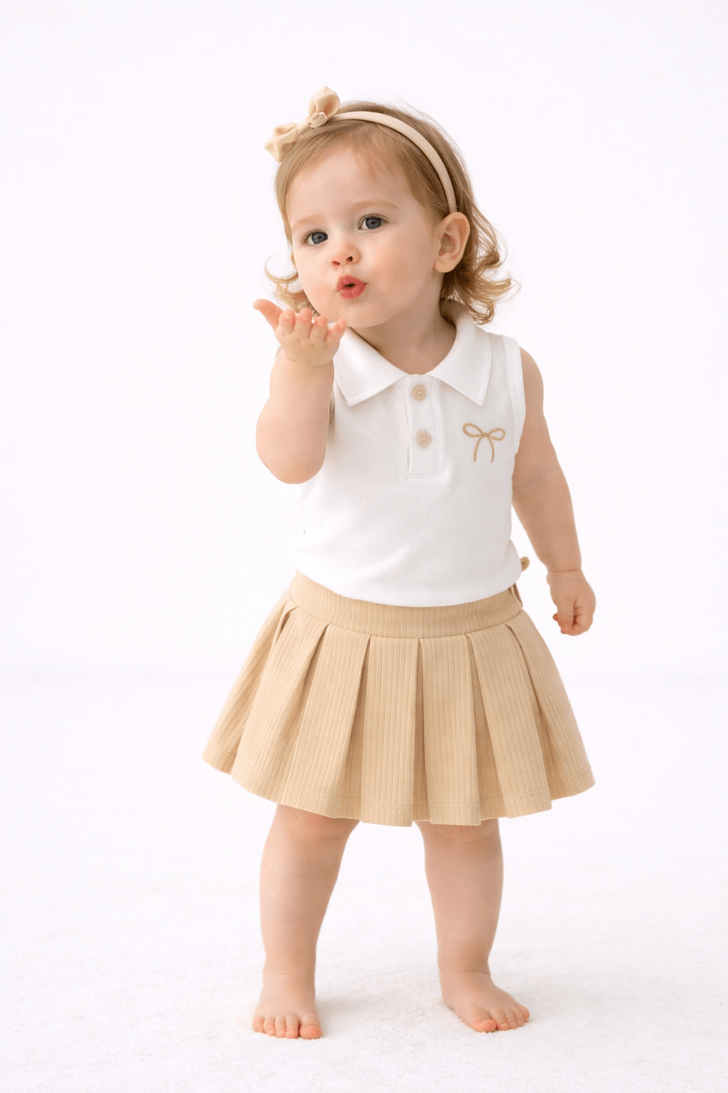 Bej pileli etek ve beyaz yakalı üstten oluşan kız bebek takım, saç bandı ile birlikte askıda sade arka plan üzerinde